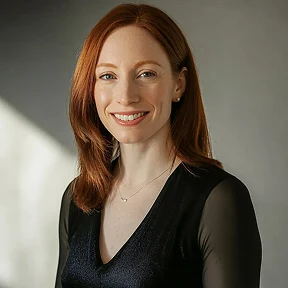 Smiling woman with red hair wearing a black top and delicate necklace against a soft gray background.